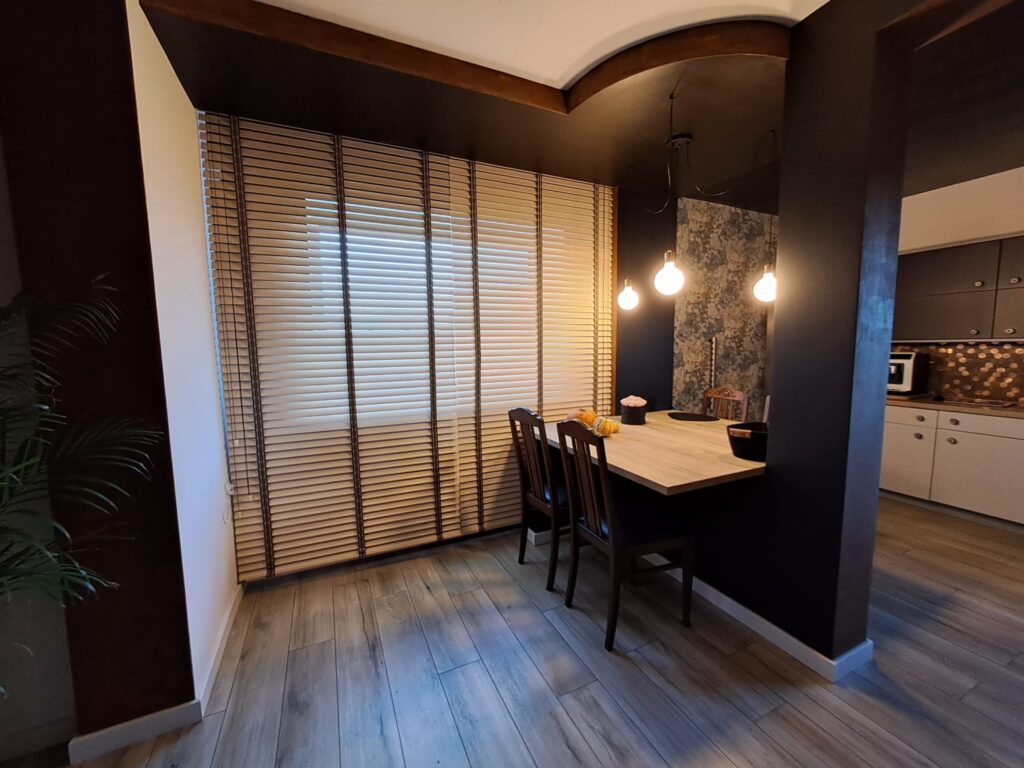 Drewniane żaluzje w jadalni z ciemnymi ścianami i trzema zwisającymi żarówkami nad stołem. Widoczne połączenie kuchni z jadalnią w nowoczesnym stylu.