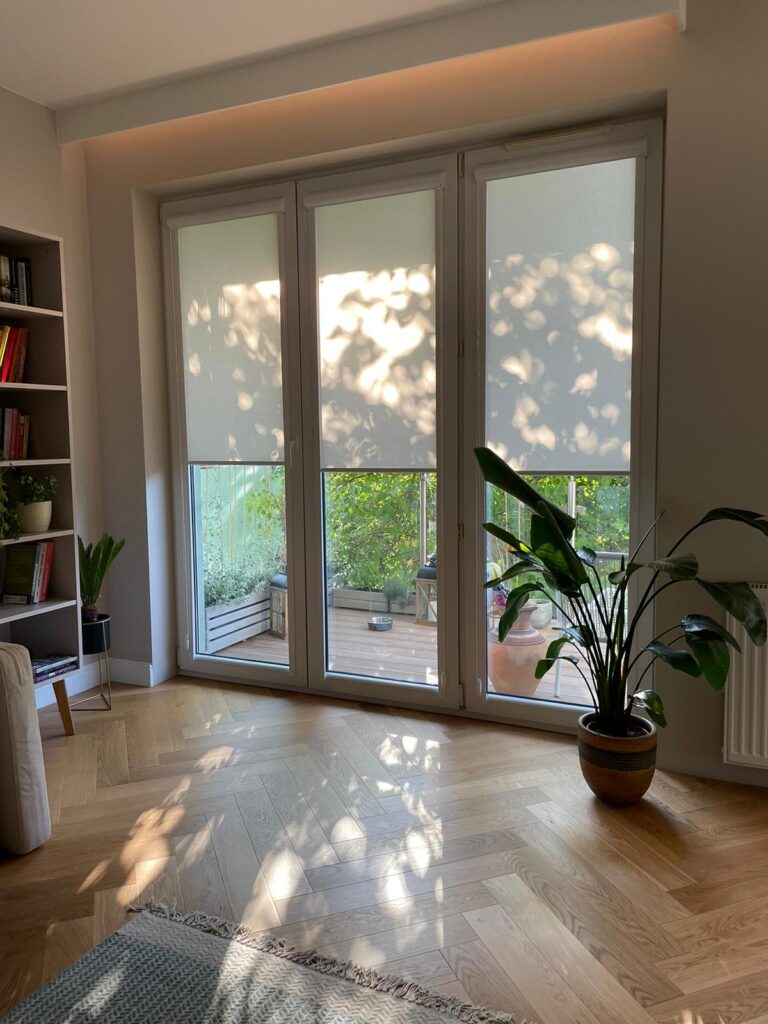 Trzy białe rolety materiałowe w dużym oknie tarasowym, filtrujące światło słoneczne wpadające do jasnego salonu z drewnianą podłogą i dużą rośliną doniczkową.
