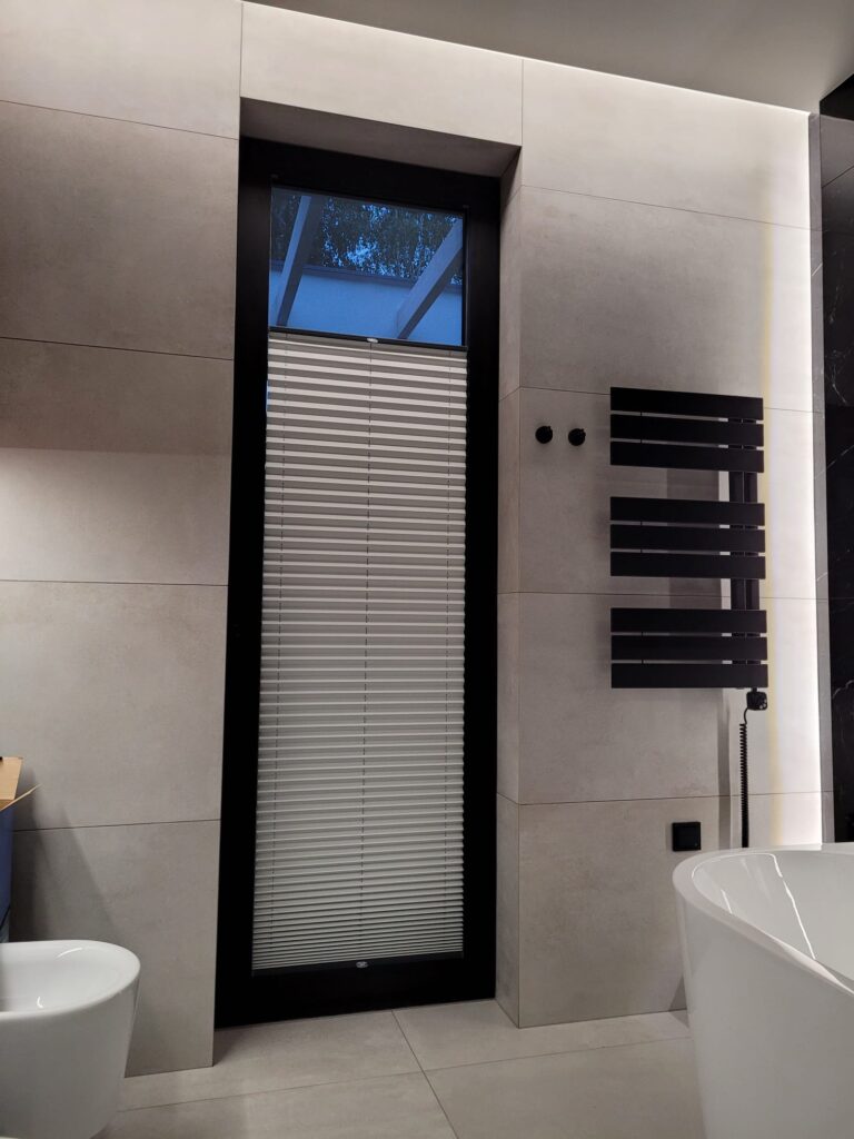 Nowoczesna łazienka z wysokim wąskim oknem zasłoniętym plisą materiałową w kolorze białym. Widoczny czarny grzejnik drabinkowy i wolnostojąca wanna.