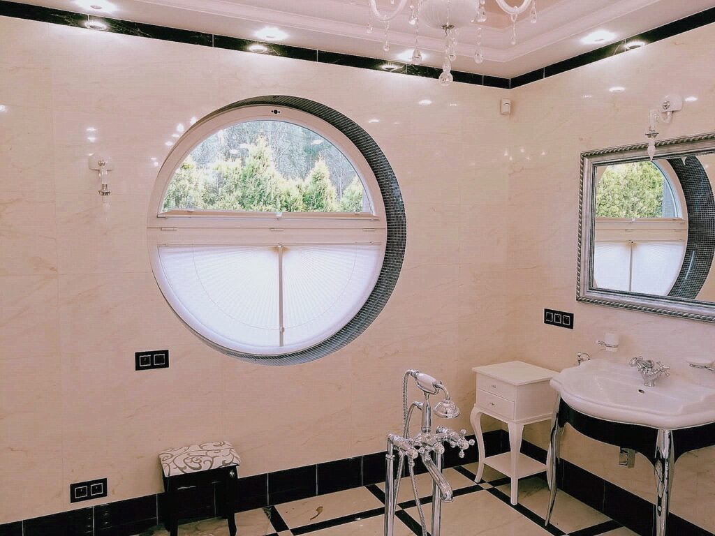 Elegancka łazienka z okrągłym oknem wyposażonym w plisy materiałowe, które zasłaniają dolną część szyby. Wnętrze w jasnym marmurze z czarnymi akcentami.