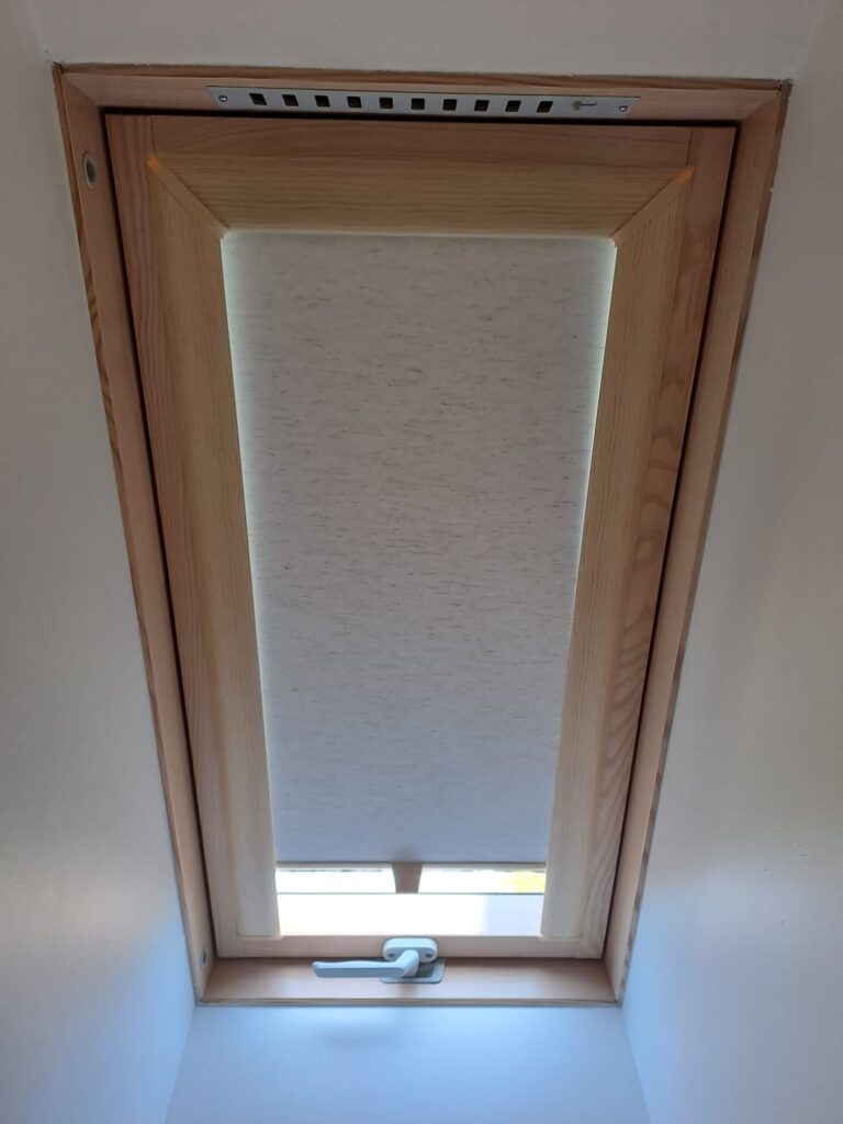 Okno dachowe z zamontowaną roletą materiałową w jasnym kolorze, zamknięte w drewnianej ramie. Widok z wnętrza poddasza, z widocznym uchwytem do otwierania.