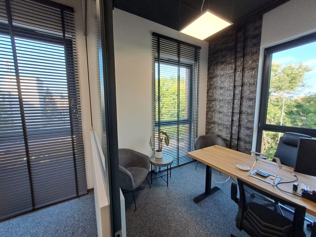 Nowoczesne biuro z dużymi oknami, na których zamontowano żaluzje aluminiowe w kolorze czarnym. Wnętrze z drewnianym biurkiem, krzesłami i industrialnym wystrojem.