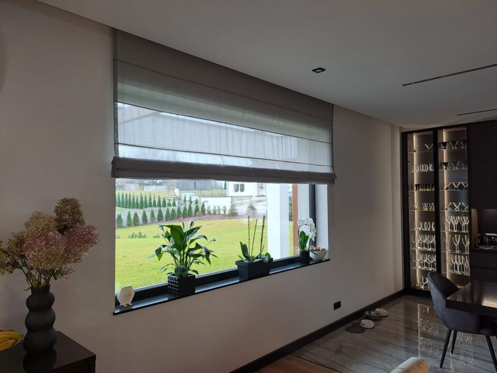 Nowoczesne wnętrze z dużym oknem ozdobionym roletą rzymską w odcieniu szarości. Na parapecie stoją doniczki z roślinami, w tle widać ogród i zabudowę jednorodzinną.