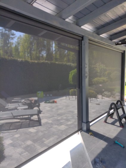 Rolety typu screen zamontowane na konstrukcji tarasu zadaszonego, z widokiem na ogród i leżaki. Materiał półprzezroczysty zatrzymujący słońce, ale pozwalający na widoczność otoczenia.