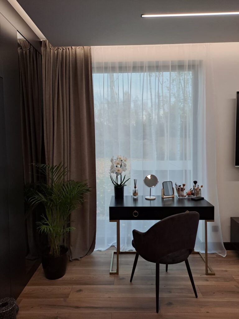 Nowoczesne wnętrze z dużym oknem zasłoniętym jasną firaną i cięższą zasłoną w kolorze taupe. Przed oknem znajduje się czarne biurko z lustrami i krzesło tapicerowane.