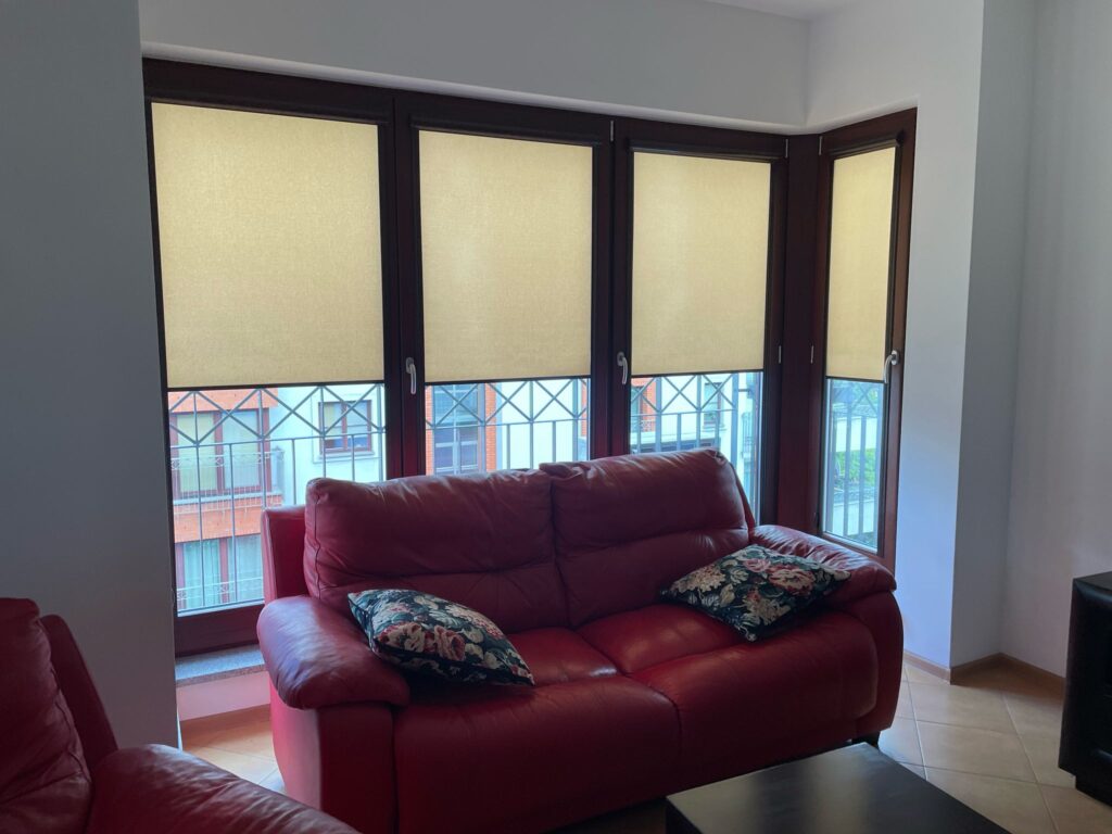 Salon z czerwonymi skórzanymi sofami oraz narożnym oknem, na którym zamontowano beżowe rolety materiałowe częściowo opuszczone. Za oknem widok na budynki mieszkalne.