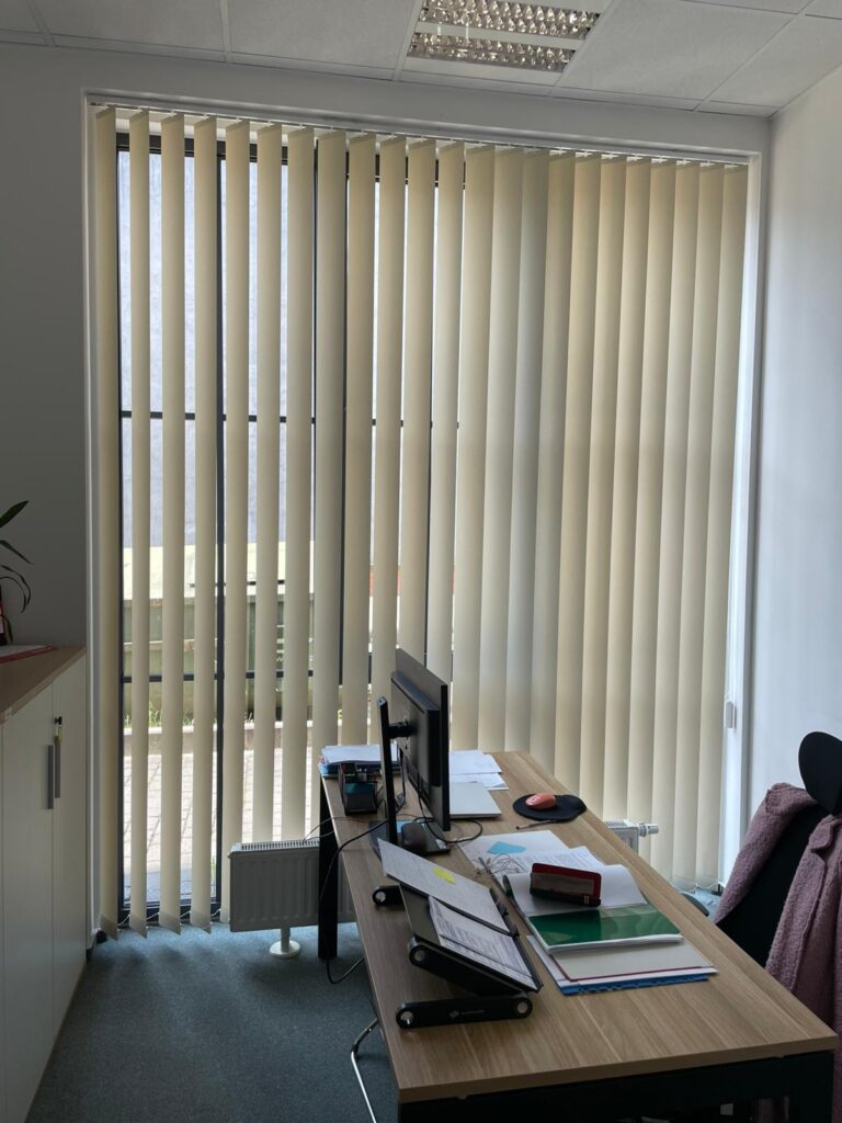 Nowoczesne biuro z dużym oknem przeszklonym, zasłoniętym jasnymi żaluzjami pionowymi. Na pierwszym planie drewniane biurko z komputerem i dokumentami.