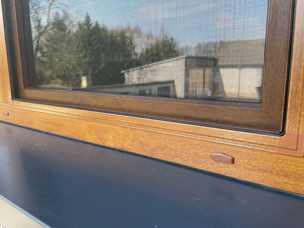 Okno z zamontowaną moskitierą ramkową w kolorze brązowym. Za szybą widoczna biała firanka i odbicie podwórka z samochodami i budynkami gospodarczo-magazynowymi.