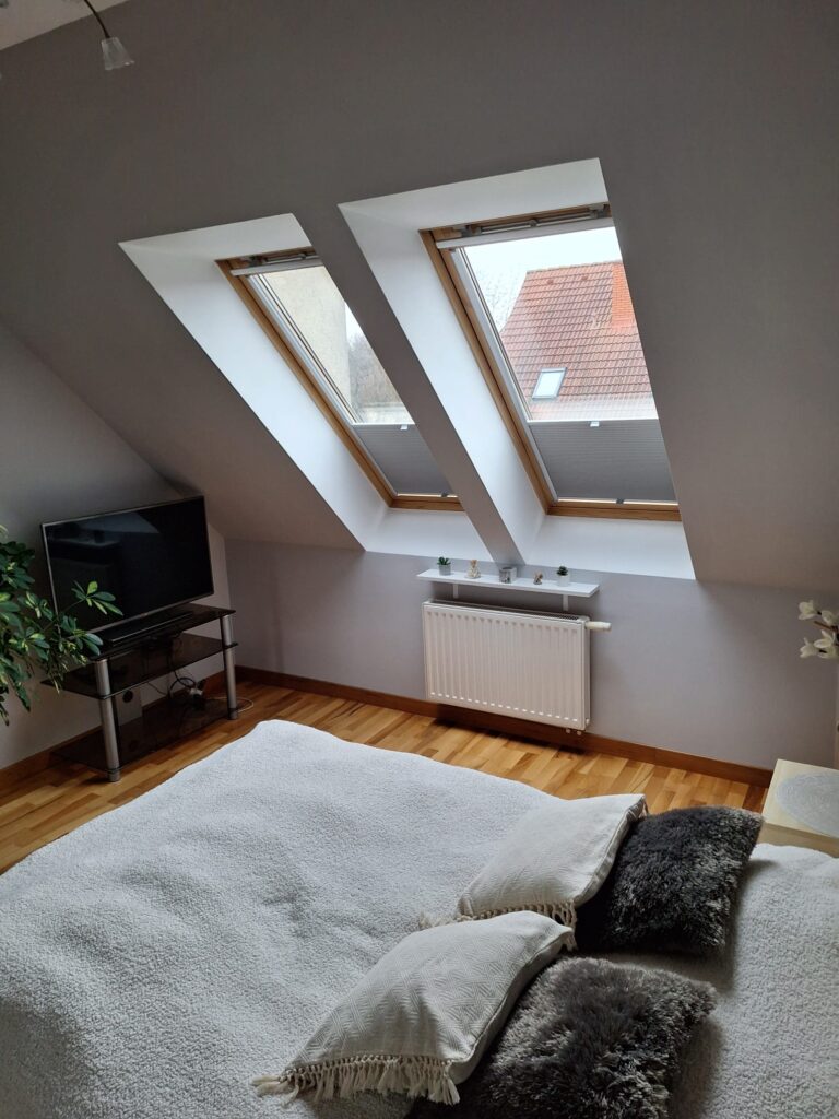 Sypialnia na poddaszu z dwoma oknami dachowymi wyposażonymi w plisy w kolorze jasnoszarym. Wnętrze z białą pościelą, dekoracyjnymi poduszkami i drewnianą podłogą.