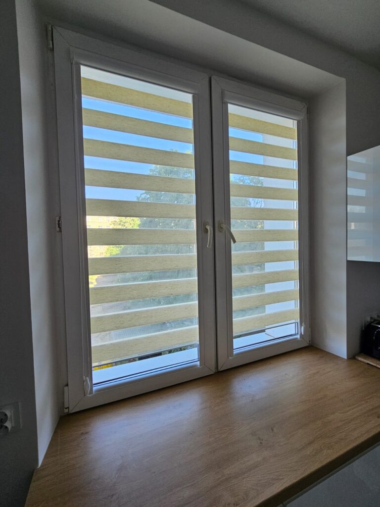 Okno kuchenne z zamontowanymi roletami dzień-noc w odcieniu jasnego beżu. Na zewnątrz widoczne zielone drzewa i ogródek. Pod oknem blat kuchenny z jasnego drewna.