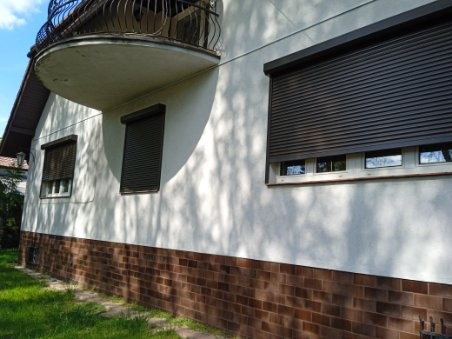 Fasada domu jednorodzinnego z zamontowanymi roletami zewnętrznymi w kolorze ciemnobrązowym. Widoczne trzy okna z opuszczonymi roletami aluminiowymi.